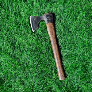 May include: A small axe with a dark metal head and a wooden handle. The axe is set against a backdrop of green grass.