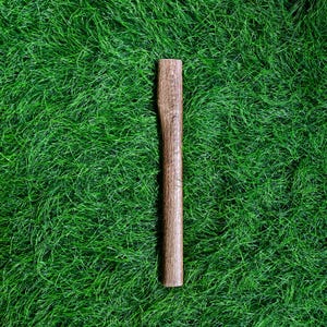 May include: A wooden handle, likely for a tool, is displayed against a backdrop of vibrant green grass. The handle is a warm brown color, with visible wood grain and a slightly curved shape for grip. The handle is approximately 10 inches long.