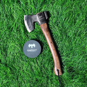 May include: A small axe with a dark metal head and a wooden handle, on green grass. The axe has a curved blade and a dark brown handle. A round, dark grey coaster with the text "DWFKHT" is next to the axe.