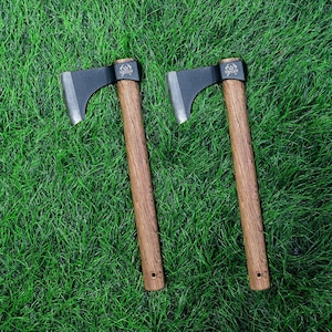 May include: Two identical throwing axes with matte black metal heads and polished silver edges. Each axe features a straight, textured brown wooden handle with a small circular hole at the base. The metal head displays a small logo with the text AXE CLUB.