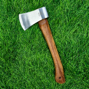 May include: A hand axe featuring a polished silver-colored metal head with a stamped logo and a curved brown wooden handle with a visible grain pattern and a small circular hole at the base of the handle for hanging or attachment purposes.