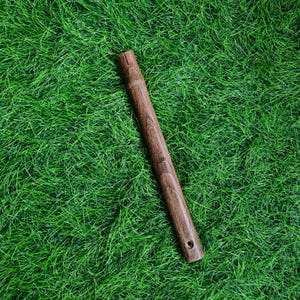May include: A long, cylindrical wooden object with a dark brown finish, featuring a carved top and a small hole at the bottom. The object is set against a backdrop of vibrant green grass.