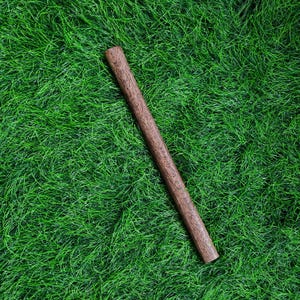 May include: A cylindrical, wooden rolling pin with a dark brown finish, resting diagonally on a bed of vibrant green artificial grass. The rolling pin's surface shows visible wood grain.