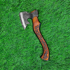 May include: A handcrafted axe with a dark metal blade and a wooden handle. The handle features intricate carved designs. The axe is set against a backdrop of vibrant green grass, showcasing its rustic appeal.