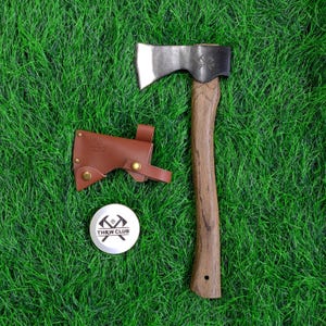 Può includere: Ascia con manico in legno e lama argentata, accompagnata da un fodero in pelle marrone e un bottone bianco THRW CLUB. L'ascia è progettata per il lancio ed è ambientata su uno sfondo di erba verde.