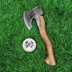 Puede incluir: Un hacha de mano con una cabeza de metal oscuro y desgastado y un mango de madera curvado, colocada junto a una ficha blanca circular que presenta un gráfico negro de dos hachas cruzadas y el texto THRW CLUB impreso en el centro.