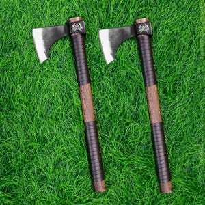 Könnte beinhalten: Zwei Äxte mit schwarzen Klingen und Holzgriffen, die mit dunkelbraunem Leder umwickelt sind. Die Axtköpfe sind silberfarben und die Griffe haben eine dunkelbraune Holzmaserung. Die Äxte liegen auf einer grünen Grasfläche.