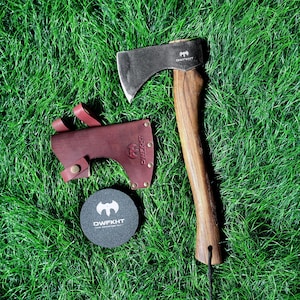 May include: Axe with a wooden handle and a dark metal blade, resting on green grass. A burgundy leather sheath and a black round coaster with white text and a logo are also present. The axe blade has the text "DWFKHT".