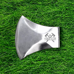 May include: A polished metal axe head with a curved blade and a flat poll. The axe head has the text "THRW CLUB" and a logo etched on the poll. The axe head is set against a background of green grass.