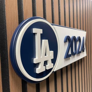 Puede incluir: Un letrero azul, blanco y color madera con las letras "LA" en blanco dentro de un círculo azul y el número "2024" en azul. El letrero está montado sobre un fondo de madera.