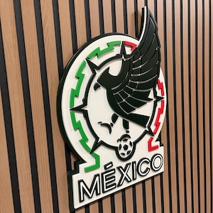 Peut inclure: Un emblème tridimensionnel de l'équipe nationale de football mexicaine, avec un cercle blanc avec des accents verts et rouges, un aigle noir et le mot "MÉXICO" en noir. L'emblème est monté sur un mur en bois.