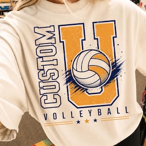 May include: A cream-colored crewneck sweatshirt featuring a large graphic print. The design includes the text "CUSTOM" vertically, a large letter "U" in gold with a blue outline, a volleyball illustration, and the word "VOLLEYBALL" below.