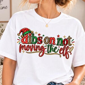 Peut inclure: Un t-shirt blanc à manches courtes avec l'inscription "dibs on not moving the elf" en rouge, vert et imprimé léopard. Un petit bonnet d'elfe rouge et vert avec des grelots dorés surmonte la lettre d. De petites étoiles ornent le motif.