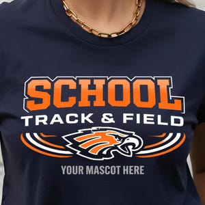 May include: Navy blue t-shirt with the words "SCHOOL TRACK & FIELD" in orange and white lettering. Below the text is a graphic of an eagle in orange and white. The text "YOUR MASCOT HERE" is below the eagle graphic.