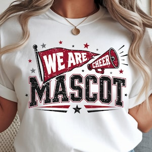 Peut inclure: Un t-shirt blanc avec un motif graphique comprenant un fanion rouge, un mégaphone rouge et le texte "WE ARE CHEER" en blanc. En dessous, le mot "MASCOT" est imprimé en grandes lettres capitales noires usées avec des contours rouges.