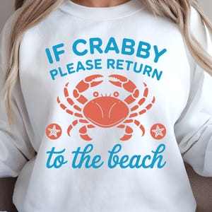 Peut inclure: Sweat-shirt blanc avec un motif de crabe orange et le texte bleu "If Crabby Please Return to the Beach". Deux étoiles de mer bleues sont de chaque côté du crabe.