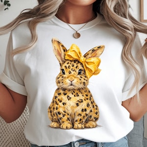 May include: White t-shirt featuring a watercolor illustration of a bunny with leopard print fur and a yellow bow. The bunny is sitting upright. The shirt has short sleeves and a round neckline.