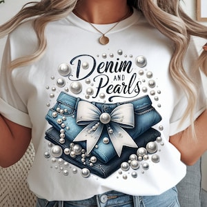 Puede incluir: Camiseta blanca con un diseño gráfico que dice "Denim and Pearls". El diseño incluye una pila de jeans azules, un lazo blanco y detalles de perlas. Se ve un collar dorado.