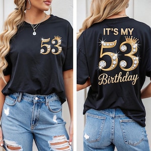 Peut inclure: T-shirt noir avec un motif doré et strass "53" et une couronne. Le dos du t-shirt porte l'inscription "IT'S MY 53 Birthday". Le t-shirt est porté avec un jean bleu. Le design est doré.