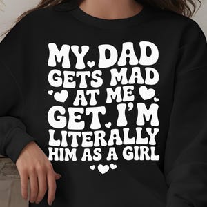 Peut inclure: Sweat-shirt noir avec l'inscription blanche "MY DAD GETS MAD AT ME. GET IT. I'M LITERALLY HIM AS A GIRL" dans une police rétro, ornée de cœurs. Vêtement décontracté.