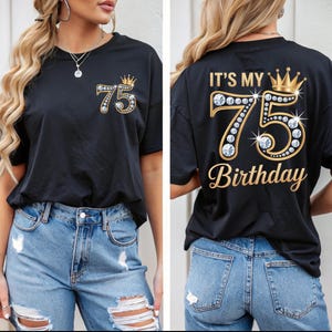 Puede incluir: Camiseta negra con un diseño dorado y pedrería "75" y una corona. La parte delantera de la camiseta presenta el número "75" con una corona. La parte trasera de la camiseta dice "IT'S MY 75 Birthday".