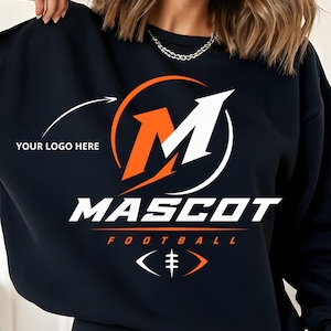 Peut inclure: Sweat-shirt noir avec le texte "YOUR LOGO HERE" et un grand logo "M" orange et blanc. En dessous, le mot "MASCOT" est affiché en blanc, avec "FOOTBALL" en dessous. Un graphisme de football est en bas.