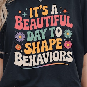 May include: Black t-shirt with the text "IT'S A BEAUTIFUL DAY TO SHAPE BEHAVIORS" in colorful, retro-style lettering. The design includes flowers and stars, creating a cheerful and educational theme.