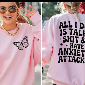 Könnte beinhalten: Hellrosa Sweatshirt mit Schmetterlingsgrafik auf der Vorderseite. Auf der Rückseite des Sweatshirts steht in schwarzer Schrift "ALL I DO IS TALK SHIT & HAVE ANXIETY ATTACKS". Das Sweatshirt hat lange Ärmel und einen Rundhalsausschnitt.