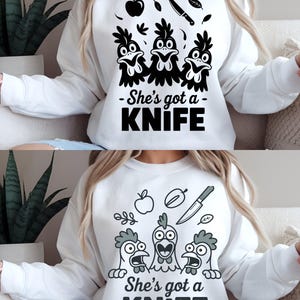 Op de afbeelding: Witte sweatshirt met een zwarte afbeelding van drie cartoonkippen en de tekst "She's got a KNIFE". Boven de kippen zijn illustraties van een appel, mes en bladeren. Een humoristisch, casual ontwerp.