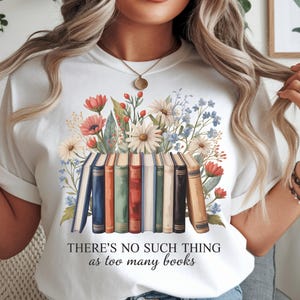 May include: White t-shirt featuring a watercolor design of books and flowers. The text "THERE'S NO SUCH THING as too many books" is printed below the illustration. The shirt has rolled-up sleeves.