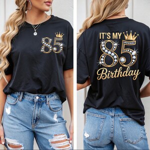 Op de afbeelding: Zwart T-shirt met een gouden en strass ontwerp van het nummer 85 en een kroon. De voorkant van het shirt heeft het nummer 85 en een kroon. De achterkant van het shirt zegt "IT'S MY 85 BIRTHDAY" in gouden letters.