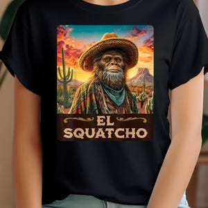 May include: Black t-shirt featuring a graphic of a Bigfoot-like creature wearing a cowboy hat and a colorful poncho, set against a desert sunset backdrop. The text "EL SQUATCHO" is displayed below the image in a brown banner.