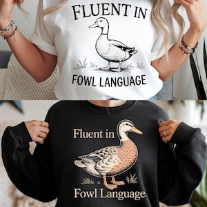 Può includere: T-shirt bianca e felpa nera con un'illustrazione di un'anatra e la scritta "Fluent in Fowl Language". L'anatra è raffigurata in uno stile cartone animato dettagliato, in piedi sull'erba. Il testo è in grassetto, carattere sans-serif.