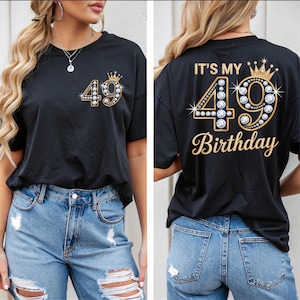 May include: Black t-shirt with a gold and rhinestone design featuring the number 49 and a crown. The front of the shirt has the number 49, and the back says "IT'S MY 49 BIRTHDAY" in gold script.