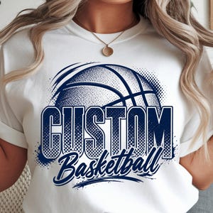Peut inclure: Un t-shirt blanc à manches courtes arborant un motif bleu foncé représentant un ballon de basket avec le texte "CUSTOM" en grandes lettres capitales et le mot "Basketball" en écriture cursive en dessous, centré sur le devant.