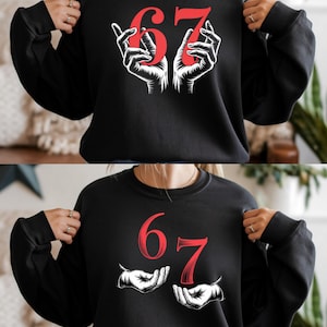 Pode incluir: Camisola preta com o número "67" em vermelho, segurado por duas mãos brancas. O desenho está centrado na frente da parte de cima de manga comprida. A camisola tem gola redonda e um corte descontraído.