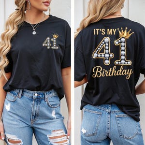 May include: Black t-shirt with the number "41" embellished with rhinestones and a gold crown. The front of the shirt has the number "41" and a crown. The back of the shirt says "IT'S MY 41st Birthday" in gold script.