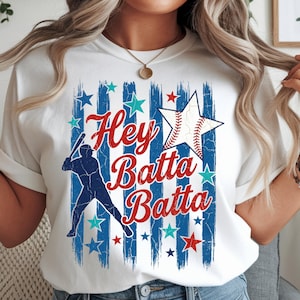 May include: White t-shirt with a baseball theme. The design features the text "Hey Batta Batta" in red script, a baseball-shaped star, a baseball player silhouette, and blue and red stars against blue stripes.