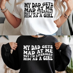 May include: Two shirts, one white and one black, each with the text "MY DAD GETS MAD AT ME BUT I'M LITERALLY HIM AS A GIRL" in a bold, retro font. The white shirt is a short-sleeved tee, and the black one is a long-sleeved sweatshirt.