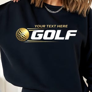 以下が含まれることがあります： 黒いスウェットシャツ。金色のゴルフボールのグラフィックと白い「GOLF」の文字。 「YOUR TEXT HERE」の文字が「GOLF」の上にあります。 ゴルフボールには点線模様があります。