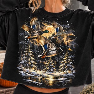 Peut inclure: Un sweat-shirt noir à manches longues avec une impression graphique représentant trois canards en vol au-dessus d'une forêt enneigée la nuit. La scène comprend une grande pleine lune, des sapins enneigés et un plan d'eau réfléchissant.