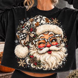 Op de afbeelding: Een zwart sweatshirt met lange mouwen en een grafische print van de Kerstman met een bril met camouflagepatroon. De Kerstman draagt een muts versierd met een zwart-witte ruitstrik, dennenappels en rode bessen, omringd door sneeuwvlokken.