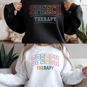 Puede incluir: Dos sudaderas, una negra y otra blanca, cada una con las palabras "SPEECH THERAPY" en letras de bloque coloridas. La sudadera negra tiene un diseño multicolor, mientras que la sudadera blanca tiene un diseño similar.