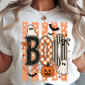 Peut inclure: Un t-shirt blanc avec une impression graphique affichant le mot BOO en grandes lettres. Le B est noir, le O est à carreaux orange et blancs, et le second O présente des rayures noires et blanches. Inclut des fantômes, une citrouille et des chauves-souris.