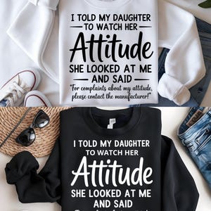 Op de afbeelding: Twee sweatshirts, één witte en één zwarte, elk met hetzelfde tekstontwerp. Het ontwerp luidt: "I TOLD MY DAUGHTER TO WATCH HER Attitude SHE LOOKED AT ME AND SAID For complaints about my attitude, please contact the manufacturer!"