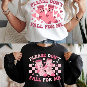 Puede incluir: Camiseta blanca y sudadera negra con el texto rosa "PLEASE DON'T FALL FOR ME". El diseño incluye dos calcetines rosas con caras, rodeados de corazones rosas. La sudadera tiene un fondo a cuadros.