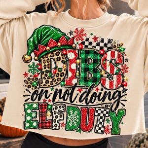 Peut inclure: Un sweat-shirt court à manches longues beige avec une impression graphique portant le texte DIBS on not doing ELF DUTY. Le motif comprend un chapeau d'elfe vert, des guirlandes lumineuses colorées, des flocons de neige et des lettres à motifs léopard, pois rouges, tartan vert et rayures rouges.
