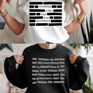 May include: White t-shirt and black sweatshirt featuring the text "everything is fine trust your government" in a block letter design. The text is in black on the white shirt and gray on the black sweatshirt. The shirts are casual wear.