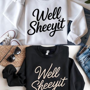 Peut inclure: Deux sweatshirts, un blanc et un noir, chacun avec l'expression "Well Sheeeyit" en écriture cursive. Le sweat-shirt blanc est associé à des baskets blanches et un jean bleu. Le sweat-shirt noir est associé à des lunettes de soleil et des chaussures marron.