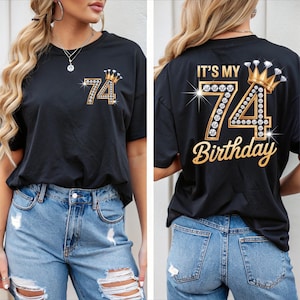 Puede incluir: Camiseta negra con un diseño dorado y pedrería con el número 74 y una corona. La parte delantera de la camiseta muestra el número 74 con una corona. La parte trasera de la camiseta dice "IT'S MY 74 BIRTHDAY" en letras doradas.